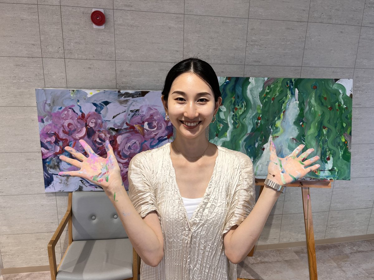 自作の絵の前で絵の具のついた両手を広げて笑うアーティストの林満里奈さん